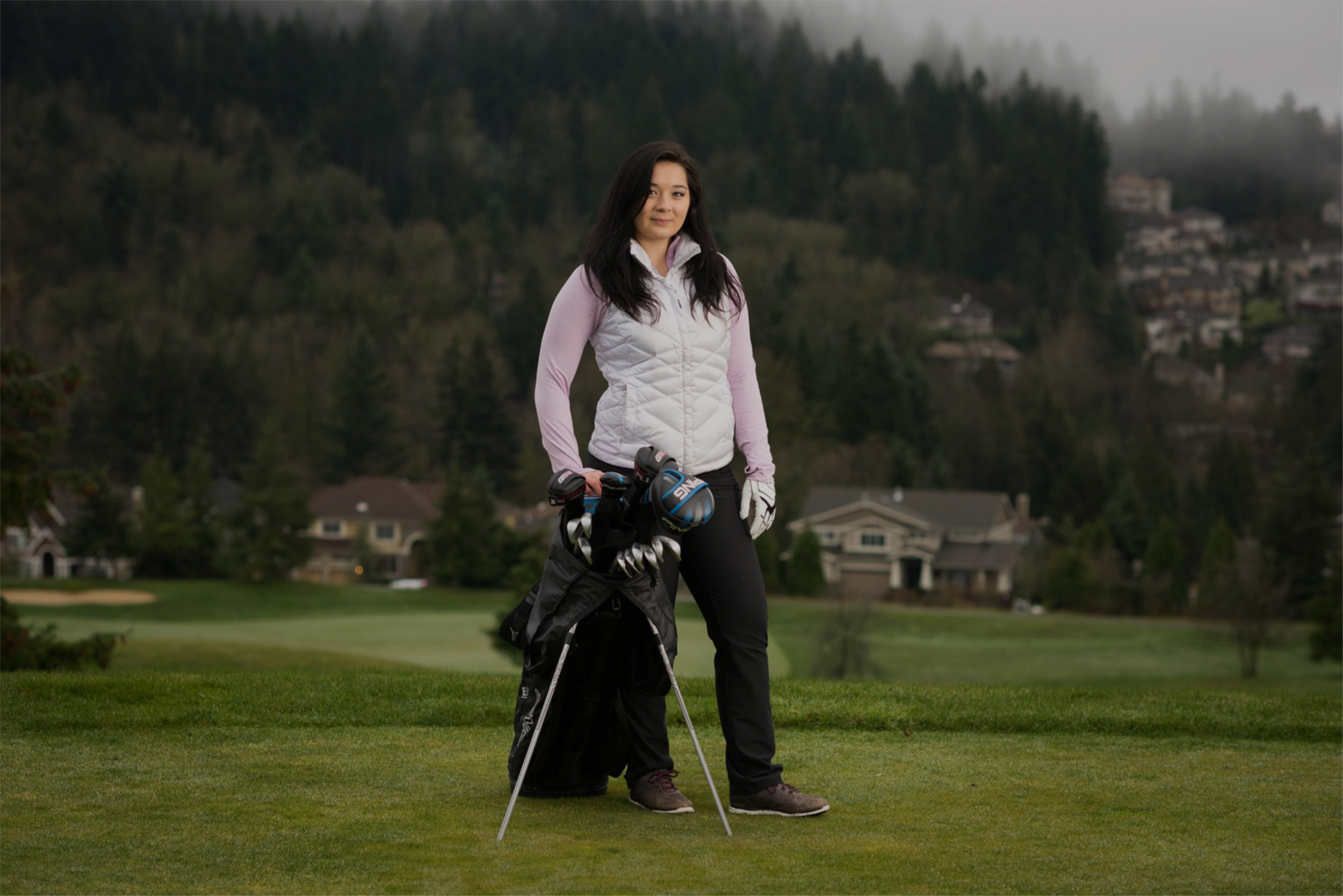 Young woman golfer posing on green with golf clubs at her side inside golf bag resting on tripod. Wearing a long sleeve and pants with brown golfing shoes and a vest jacket. Background includes, grass, trees, homes and fog.