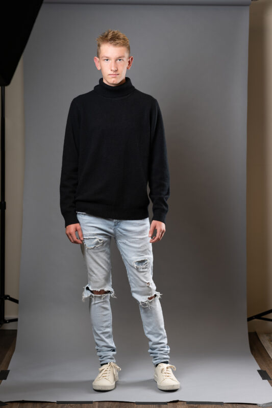 Young man posing while standing in front of a photograpers backdrop wearing light colored jeans and black long sleeve sweater.