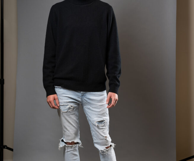 Young man posing while standing in front of a photograpers backdrop wearing light colored jeans and black long sleeve sweater.