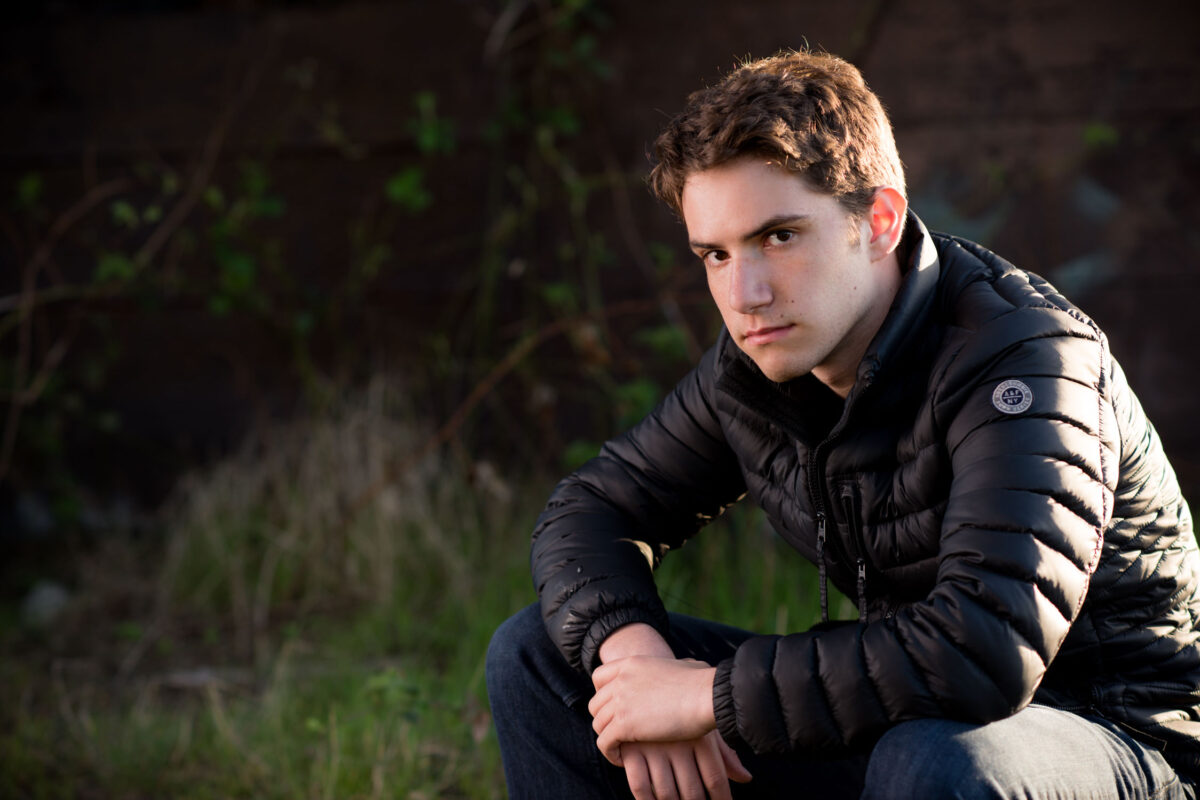 A High School Boy wearing a black down jacket while sitting close to ground and staring. Background is blurred trees and foliage.