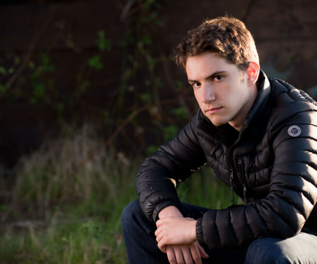A High School Boy wearing a black down jacket while sitting close to ground and staring. Background is blurred trees and foliage.