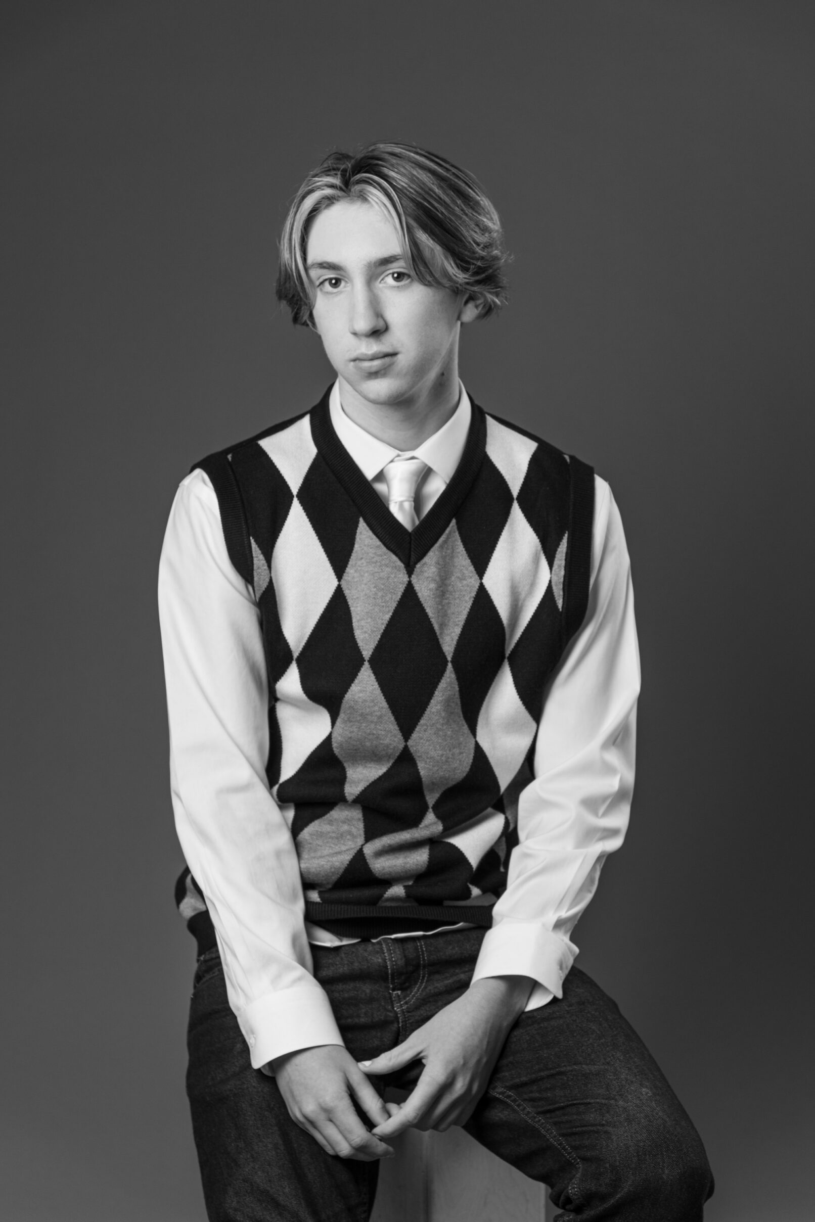 black and white portrait of a teenager wearing a sweater vest white shirt with tie and jeans posing on a stool.