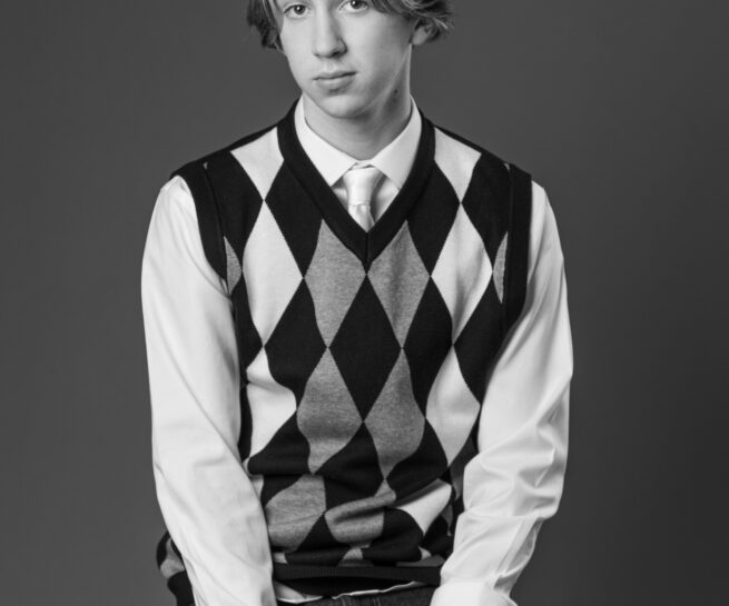black and white portrait of a teenager wearing a sweater vest white shirt with tie and jeans posing on a stool.