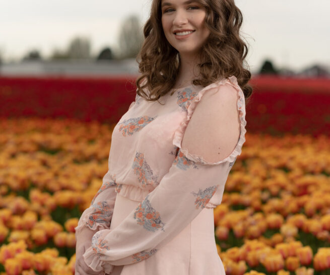 Smiling lady wearing a pant suit with tulips and blurred tree line in background.