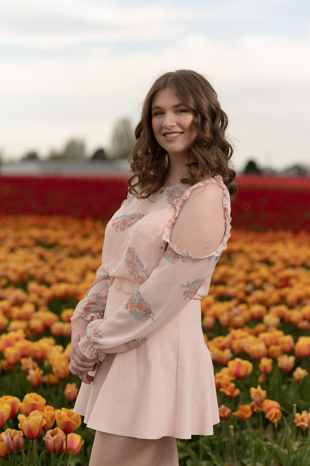 Smiling lady wearing a pant suit with tulips and blurred tree line in background.