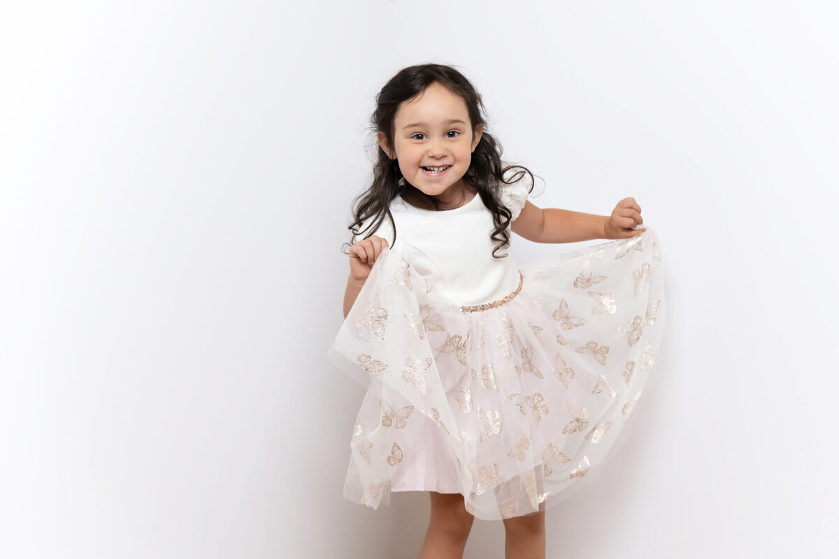 Lively portrait of preschool girl grinning and dancing in white dress with gold butterflies overlaid onto white lace.