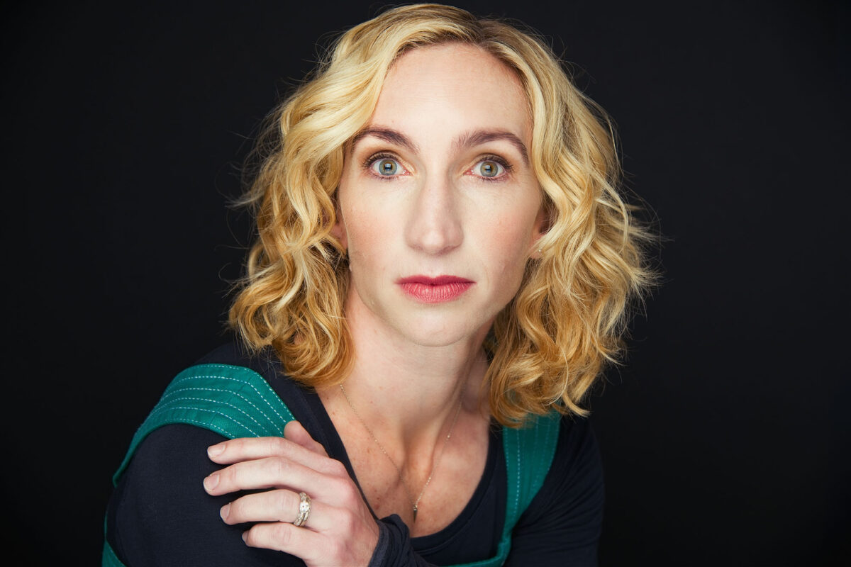 woman posing for headshot portrait. Wearing a dark colored shirt with light blue stiched overalls, blond hair, wide eyed stair with hand resting on shoulder with a diamond wedding engagement ring and wedding band.