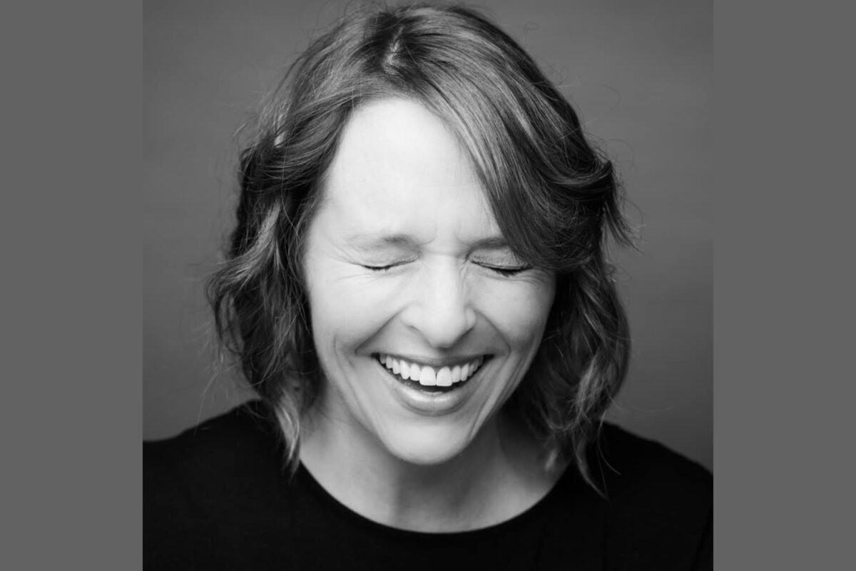 black & white headshot woman laughing with squinting closed eyes wearing a black tee shirt.