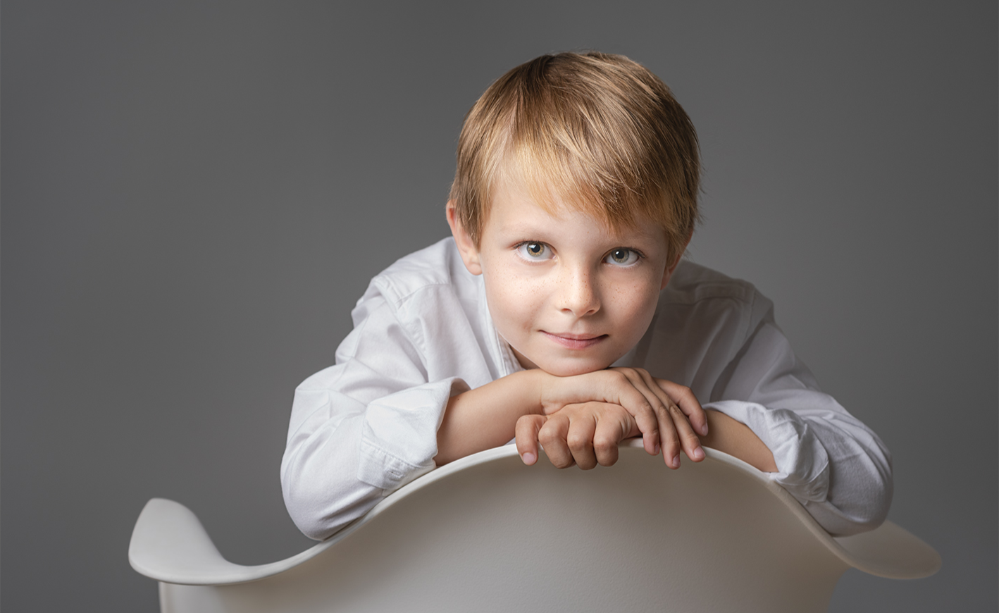 Yong man posing for portrait. Sandy blond hair, long sleeve white shirt, sitting onknees while facing camera with hands on back of chair for a casual pose.